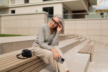 Obraz premium Senior woman sitting on outdoor steps in modern urban setting, enjoying sunny day while using smartphone