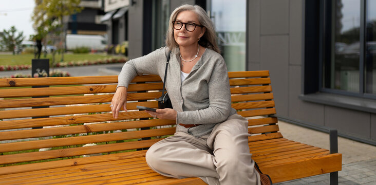 Senior woman relaxing on a wooden bench in an urban setting while using her smartphone on a sunny afternoon