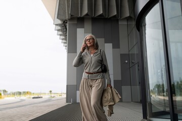 Senior woman leaving modern building while talking on smartphone in urban setting during daylight