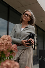 Senior woman poses confidently with a camera near blooming flowers in a modern urban setting during daytime