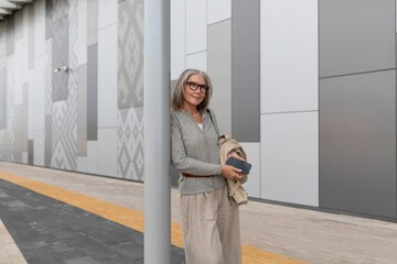Confident woman stands by modern urban wall while holding phone and jacket in late afternoon sunlight