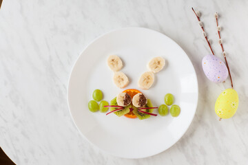 Creative healthy snack of breakfast for kids fruit bunny shaped on white plate, easter eggs around. Rabbit, basket makes from grape, orange, banana, apple. Top view