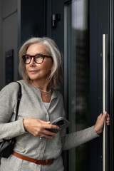 Woman with glasses steps outside a building while checking her smartphone on a sunny day