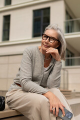 Senior woman sitting on bench outdoors, enjoying a moment of reflection in a modern urban setting with building backdrop