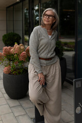 Elderly woman poses confidently outside modern building surrounded by blooming flowers in late afternoon light