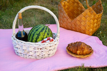 Outdoor picnic setup with a basket of fresh watermelon and pastries on a pink blanket in a grassy...