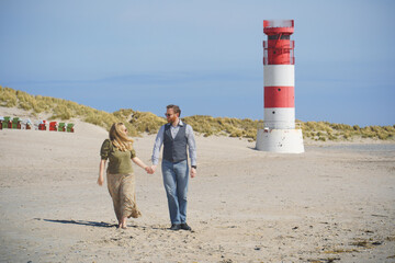 A loving couple strolls the sandy beach of Helgoland Düne with the iconic red-and-white lighthouse in the background, symbolizing romance, travel, and coastal lifestyle.