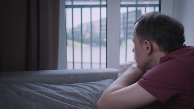 A man looks out of the window, observing the movement outside, leaning on the back of the sofa