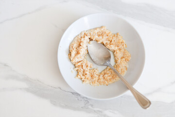 Creamy oatmeal breakfast in white bowl on marble surface with metal spoon