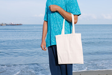 Man with black, tan skin holding in hand white eco cotton textile bag. Blue water of sea or ocean coast background. Seaside, sunny day. Blank mock up tote bag, eco concept, place for text design logo