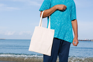 Man with black, tan skin holding in hand white eco cotton textile bag. Blue water of sea or ocean...