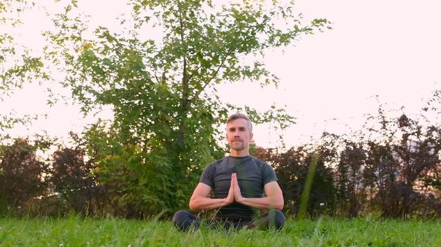 Happy middle aged man practicing yoga sitting in lotus pose on green grass in park outdoors