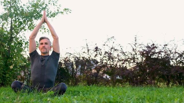Mid aged man practicing yoga sitting in lotus pose on green grass in park