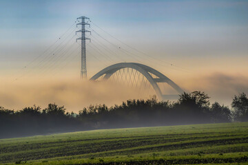 A fog-covered bridge and power line emerge in the morning landscape, evoking a mysterious autumn...