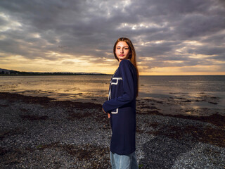 Young enthusiastic girl in stylish black coat posing on a beach at sunset, ocean and dark sky in...