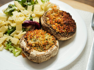 A plate of food with a fork and knife on a wooden table. The plate has a fresh salad with cheese and two fancy posh stuffed mushroom patties. Simple tasty meal.