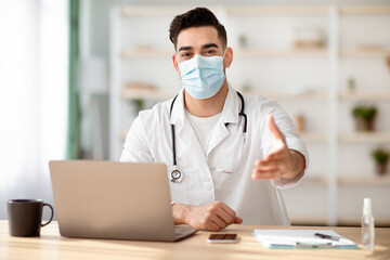 Young middle-eastern doctor in protective face mask sitting at workdesk with laptop at clinic,...