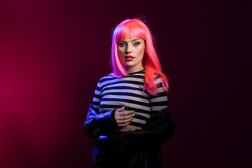 Stylish young woman in striped crop top presents unique look with intense stare in dramatic color studio. Trendy female individual stands boldly under magenta spotlight wearing edgy fashion attire.