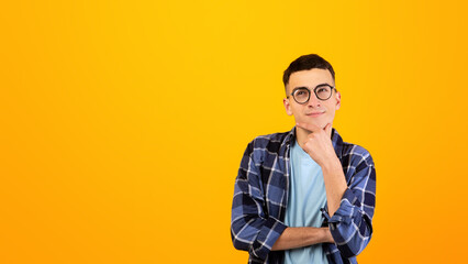 Pensive young man in glasses touching his chin, thinking over something on orange studio background. Puzzled hipster guy deep n thought, making choice or decision, imagining or daydreaming