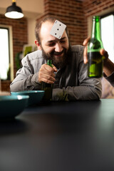 Caucasian guy relaxes with friends, smiling with card on head and beer in hand during entertaining social gathering. Young white man enjoys bonding time, playing fun guessing game at table.