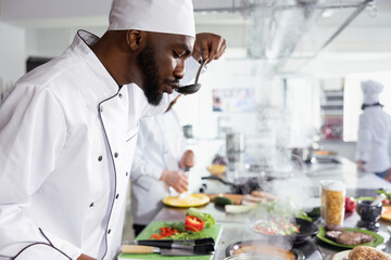 Black culinary expert tasting a pan of simmering sauce from the stovetop, making final adjustments to enhance rich and savory flavor. Chef life with delivering gourmet experiences.