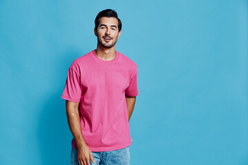 Full body cheerful smiling young man wearing pink t-shirt standing against a solid blue background,...