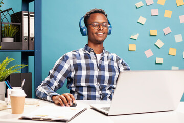 Young businessman with headphones, working on his laptop, brainstorming ideas for startup company. Entrepreneur using his mouse, scrolling and preparing project presentation on his personal computer.