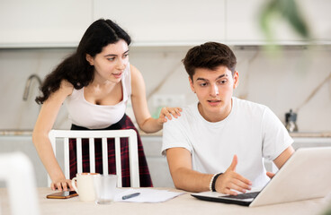 Fototapeta premium Sad and tired freelancer husband works at home on laptop and listens to her wife reproaches in frustration. Family quarrel, lack of free time, unmet needs