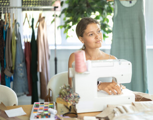 Qualified dressmaker sews clothes on a machine in a workshop. Thoughtful seamstress sitting at table and working