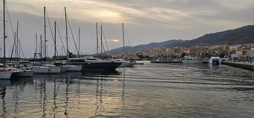 Yachts and sailboats docked in a calm marina at sunset, with city buildings and mountains in the...