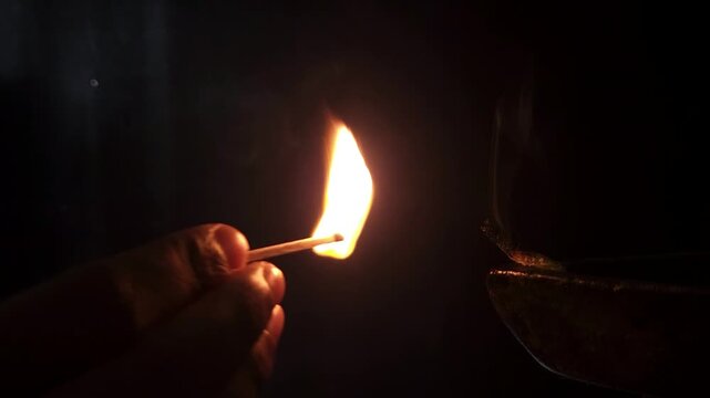 Slow-motion horizontal shot from India, a hand delicately lights a diya against a black background, capturing the essence of spiritual preparation and the vibrant warmth of Diwali festival celebration