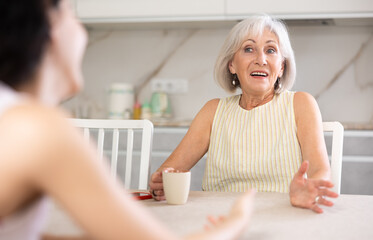Elderly mother and adult daughter drinking tea and chatting happily at home
