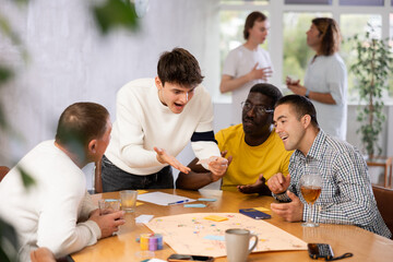 Group of enthusiastic men playing board game sitting around large table while active player is...
