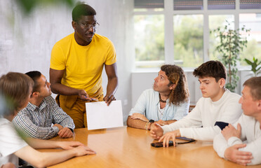 Adult African American tutor, holding sheet of paper, conducting lesson for group of interested men of different nationalities sitting at table in foreign language school classroom