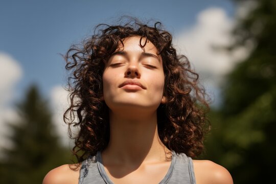 A young woman with curly hair is meditating with eyes closed in outdoor setting, bathed in sunlight, concept for wellness promotion, mindfulness practice and stress reduction