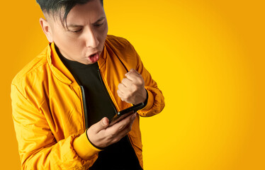 Latin man celebrating good news on smartphone with fist raised, excited reaction isolated on yellow...