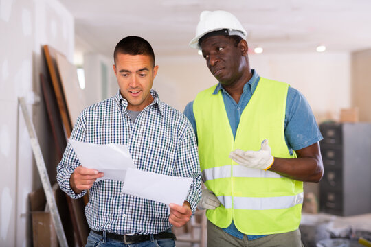 Caucasian man construction manager and african-american builder discussing project documentation in apartment during repair works.