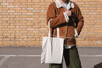Young woman holding blank white tote bag canvas fabric, city street, brick wall background. Female consumer hold big white textile shopper. template mock up design for text or logo place