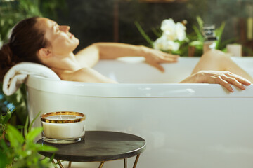 Serene side view of a relaxed woman with brown hair in a white bathtub, head on towel, hands on tubs edge. Glowing candle on table with green foliage enhances this peaceful self-care scene.