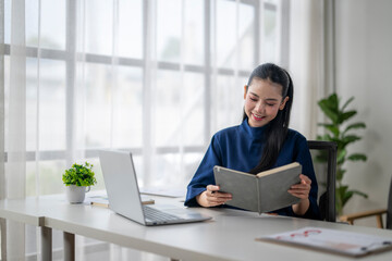 Smiling businesswoman reading book at office desk with laptop