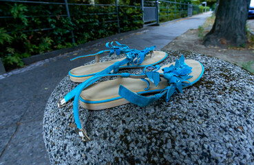 Broken blue women’s sandals left behind on a round stone post next to an empty sidewalk, shallow...