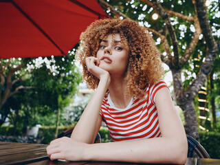 Curious young woman with curly hair wearing a red striped shirt, resting her chin on her hand...
