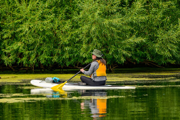 Active outdoor lifestyle: happy smiling young adult woman sitting on stand up paddleboard on a pond in summer with lush green vegetation in background