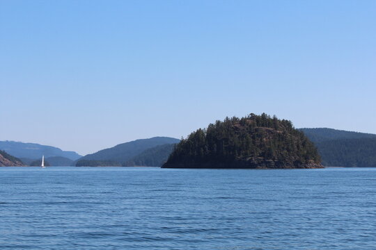 Sailing in Desolation Sound, British Columbia. A sailboat navigates calm emerald waters past rugged rocky cliffs covered with evergreen forest, under a wide blue summer sky. 