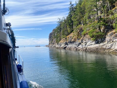 Scenic coastal view in Desolation Sound, British Columbia. A boat glides past rocky cliffs lined with evergreen trees, reflecting in calm green water under a bright blue sky with streaked clouds. 