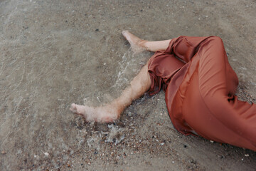 Woman's legs submerged in shallow water at the beach, wearing rust-colored pants, peaceful and calm mood with gentle waves and sandy shore.