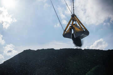 A large excavator crane is actively engaged in loading and unloading a coal bucket at a mining site. The crane's scoop is positioned above a high pile of coal, with black coal cascading down from the © Vladimir Razgulyaev