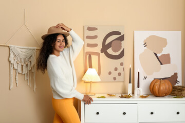 Beautiful young happy African-American woman in stylish outfit with autumn leaves, pumpkin and hat at home
