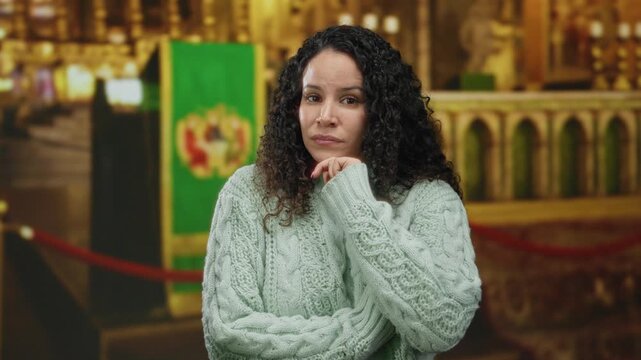 Young hispanic woman contemplating quietly indoors at a church, surrounded by holy decor, wearing a cozy sweater, capturing a moment of reflection.