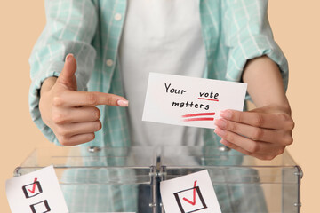 Woman pointing at paper with text YOUR VOTE MATTERS near ballot boxes on beige background, closeup